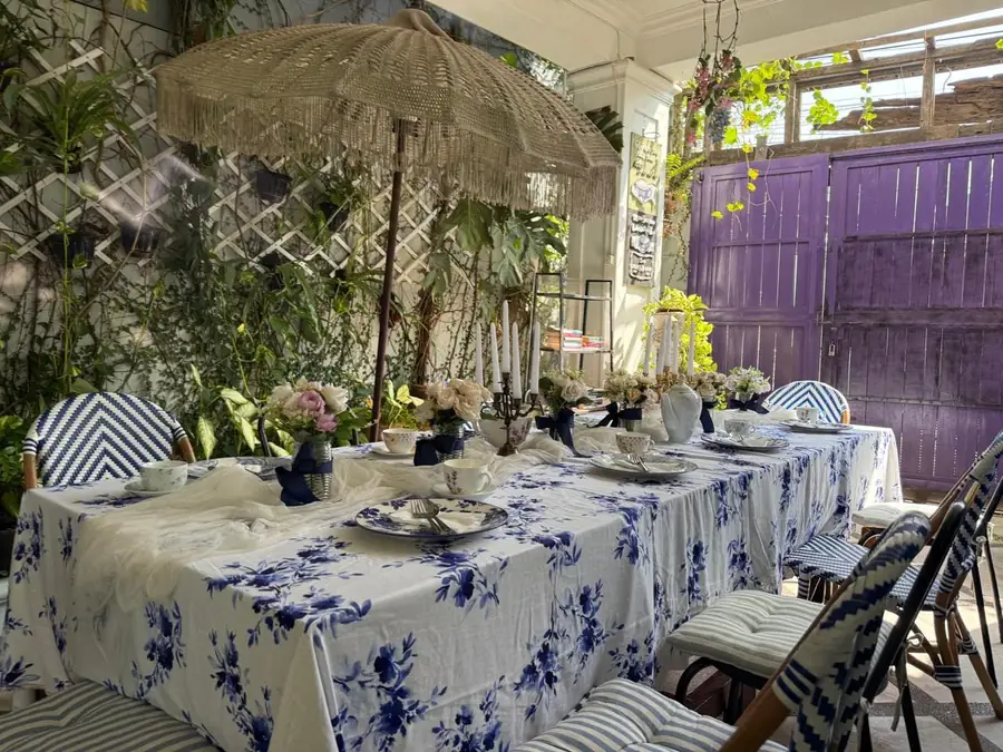Meja berkain biru putih tertata di taman dengan payung rotan